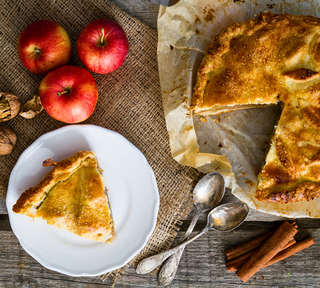 Top-down view of a rustic homemade apple pie with a slice on a plate, surrounded by fresh red apples, cinnamon sticks, and walnuts for a fall baking recipe.